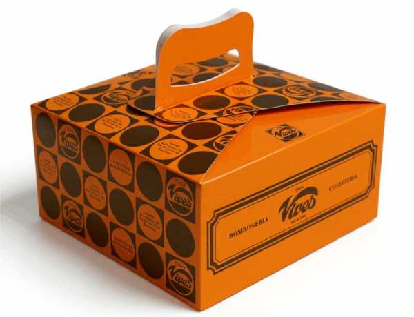 Caja de cartón naranja con asa superior, diseño gráfico retro y logotipos repetidos de una pastelería tradicional, fotografiada con iluminación profesional sobre fondo blanco